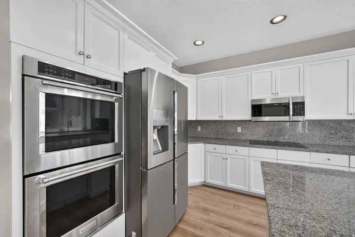 White shaker-style cabinetry, stainless steel double wall ovens, stainless steel refrigerator, stone countertops with matching backsplash, and wood-finish flooring
