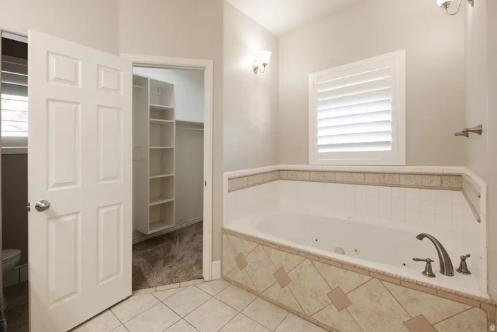 Soaking tub with tiled surround, plantation shutters, wall sconces, and built-in closet shelving
