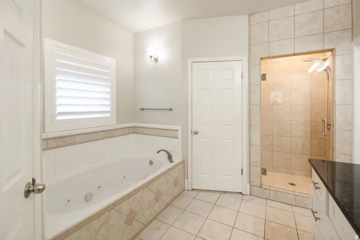 Jetted soaking tub with tile surround, walk-in shower with glass enclosure, tiled flooring, plantation shutters, and dark countertop vanity