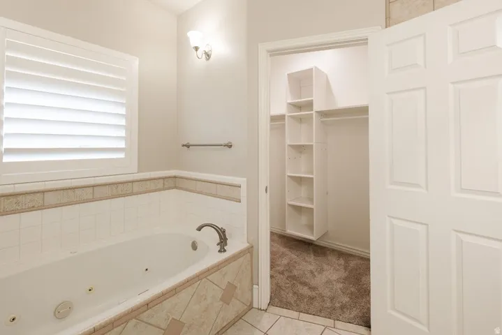 Jetted bathtub with tiled surround, plantation shutters, wall-mounted sconce, and an adjacent walk-in closet with built-in shelving