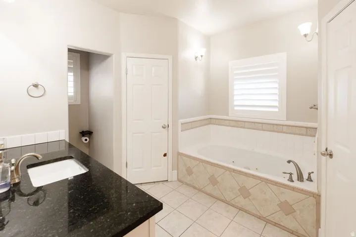 Bathroom featuring a built-in jetted tub with tile surround, a dark countertop vanity with undermount sink, sconce lighting, and white plantation shutters