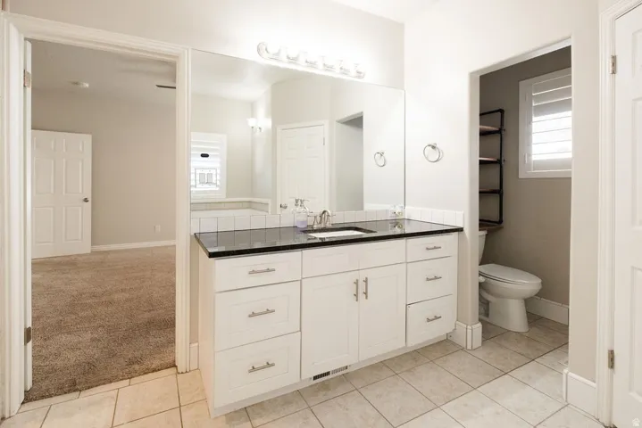 White vanity with a dark countertop and undermount sink, large wall-mounted mirror, ceramic tile flooring, and white plantation shutters