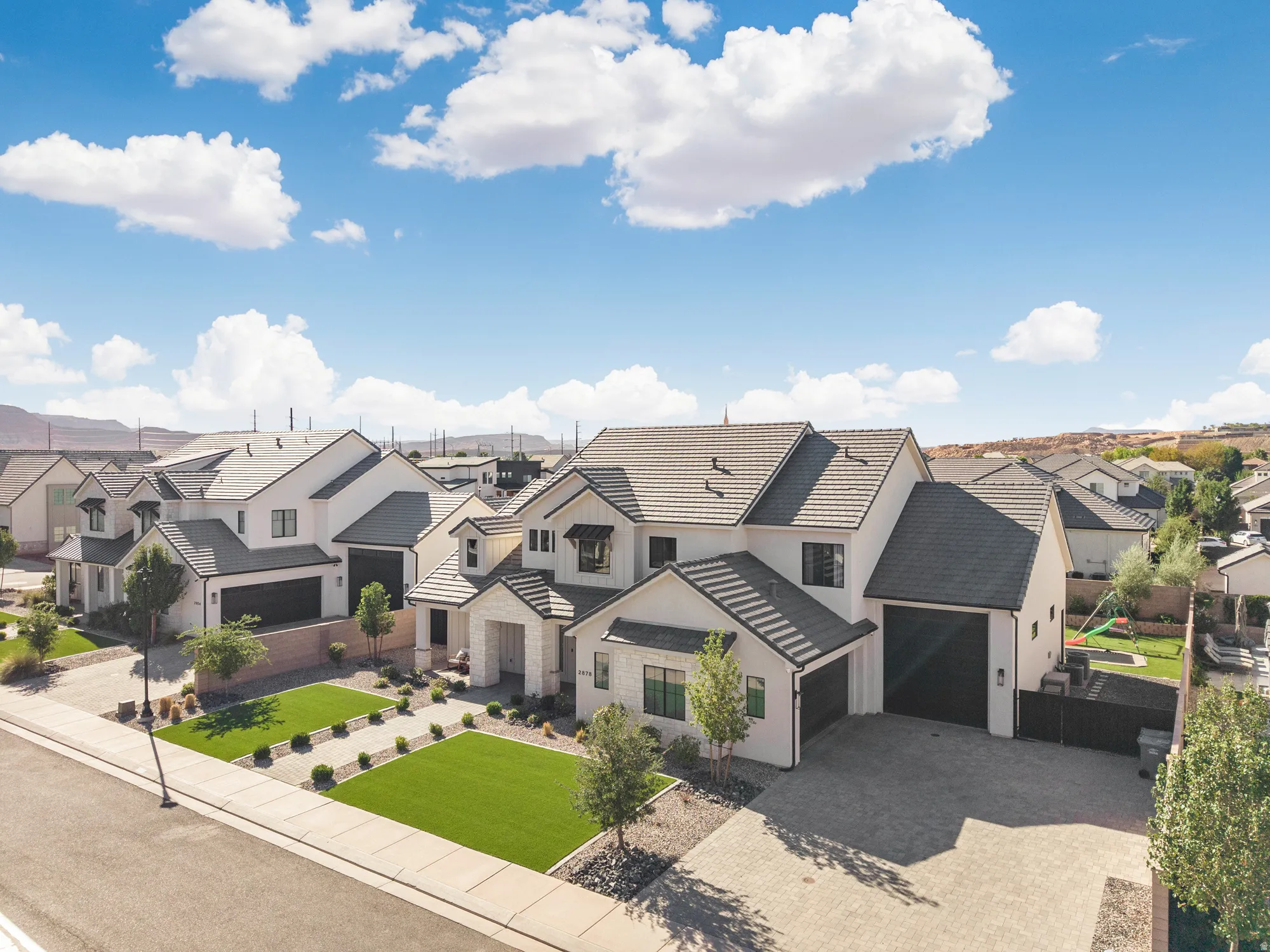 Modern exterior with a light stucco finish, dark roof tiles, an attached multi-car garage, paver driveway, and a landscaped front yard with artificial turf