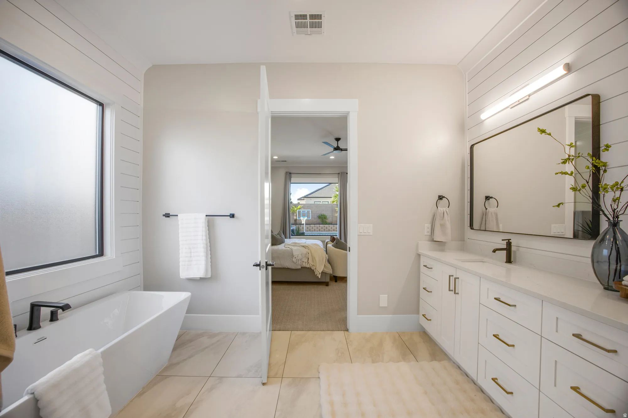 Freestanding soaking tub with black fixtures, dual vanity with white cabinetry, rectangular mirror, wall-mounted lighting, and wood-finish flooring
