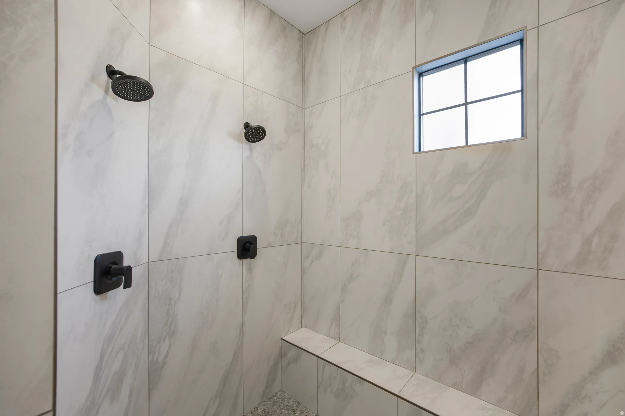 Walk-in shower with large format tile, dual matte black showerheads, built-in bench, and pebble-finish shower pan