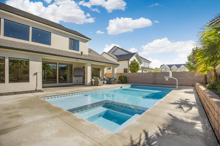 Rear exterior featuring a large swimming pool with an integrated spa, extensive concrete patio, covered patio area, and mature landscaping