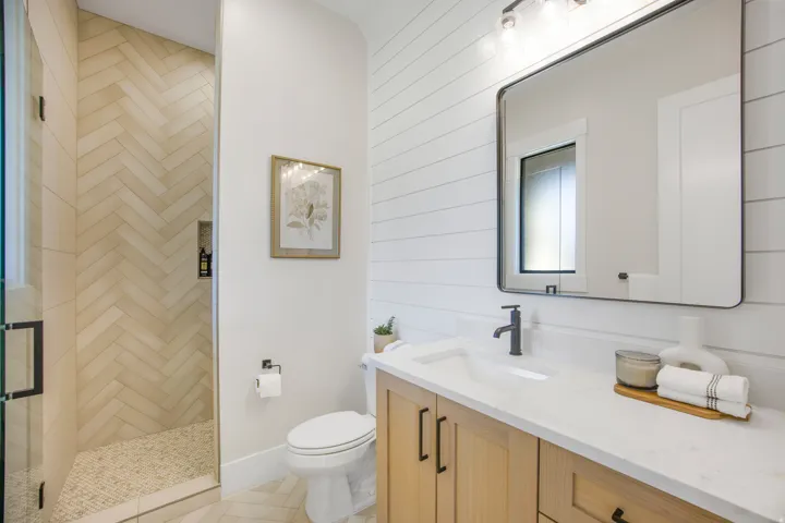 Herringbone tile shower with pebble floor, light wood-finish vanity with quartz countertop, matte black fixtures, and shiplap accent wall