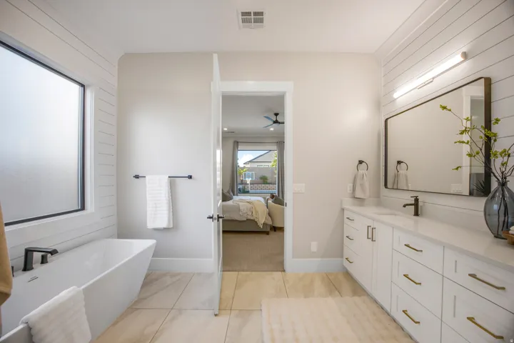 Freestanding soaking tub with black fixtures, dual vanity with white cabinetry, rectangular mirror, wall-mounted lighting, and wood-finish flooring