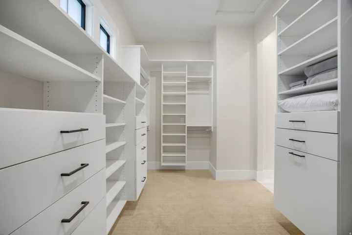 Spacious walk-in closet featuring custom white cabinetry with multiple drawers and shelving throughout, neutral wood-finish flooring, and abundant storage with organized compartments for clothing and linens