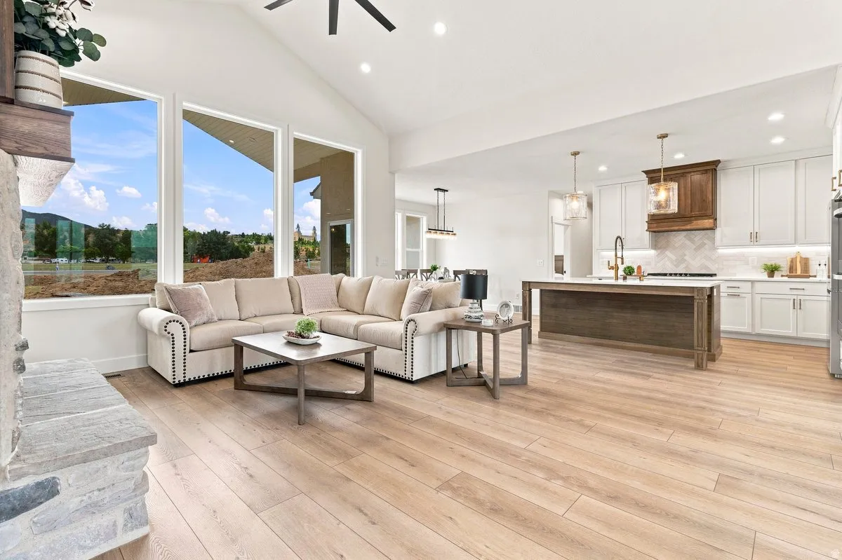Open-concept living space with cathedral ceilings, large windows, wood-finish flooring, a stone fireplace, and a kitchen featuring a central island and white cabinetry