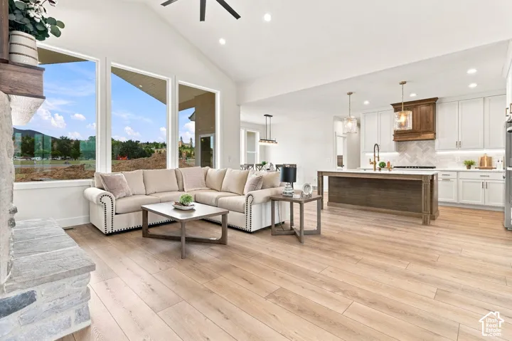 Open-concept living space with cathedral ceilings, large windows, wood-finish flooring, a stone fireplace, and a kitchen featuring a central island and white cabinetry