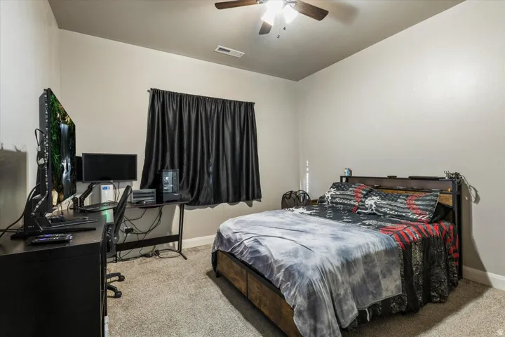 Carpeted bedroom with a wood-finish bed frame, ceiling fan, and window with dark curtains