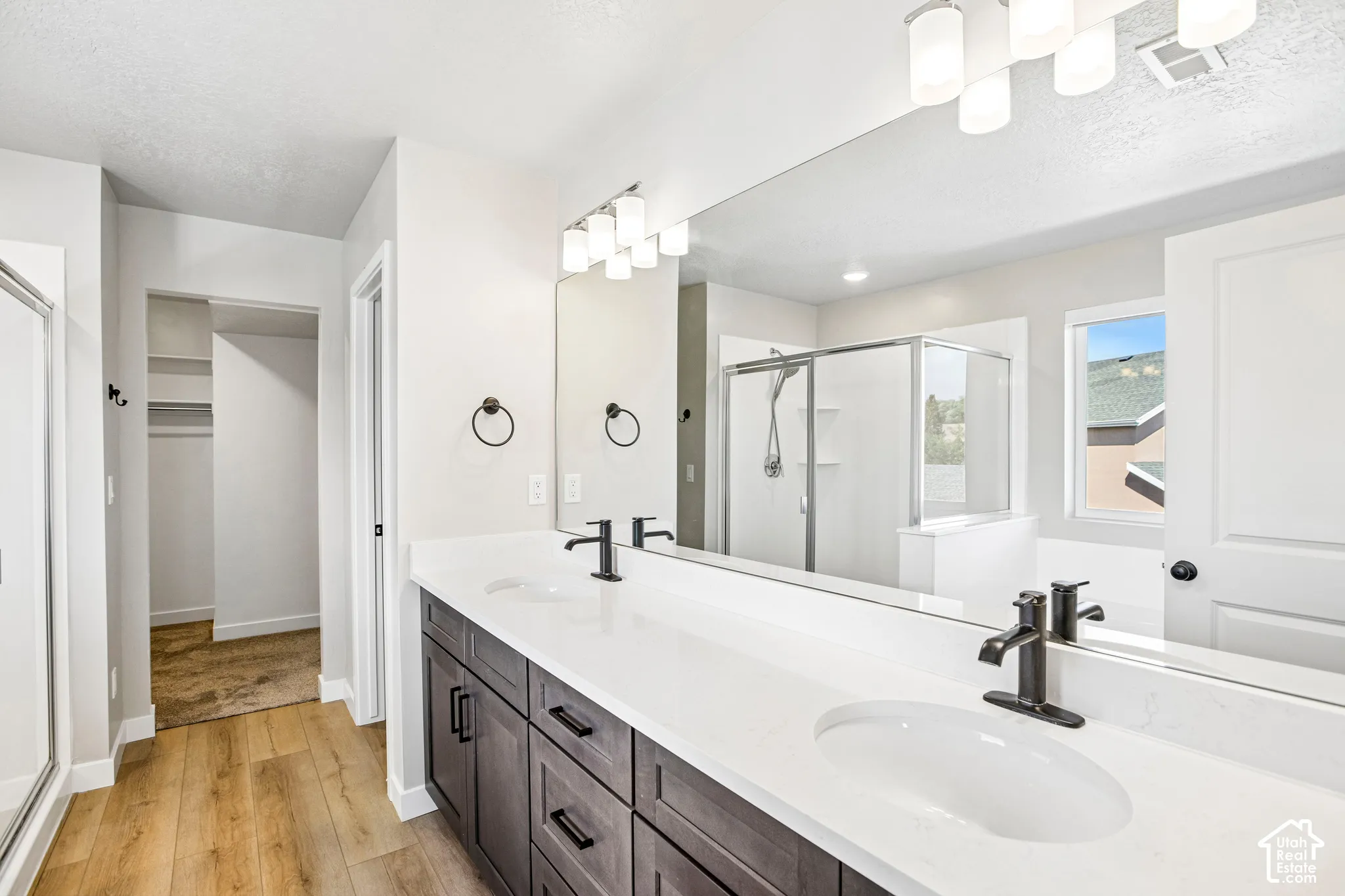 Dual vanity with white countertops, wood-finish flooring, a glass-enclosed shower, a large mirror, and recessed lighting