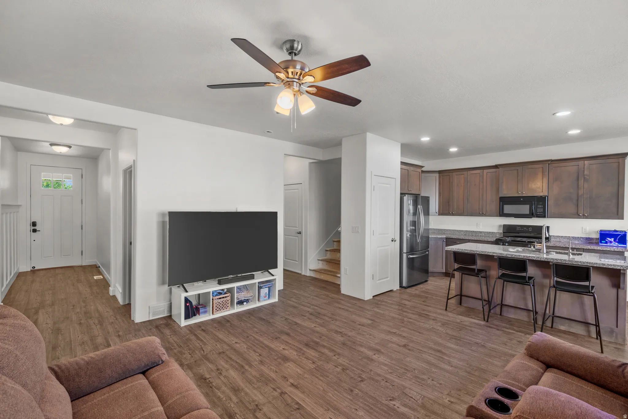 Wood-finish flooring throughout, kitchen with dark wood cabinetry, stainless steel refrigerator, and built-in microwave, kitchen island with seating, and a ceiling fan with integrated lighting