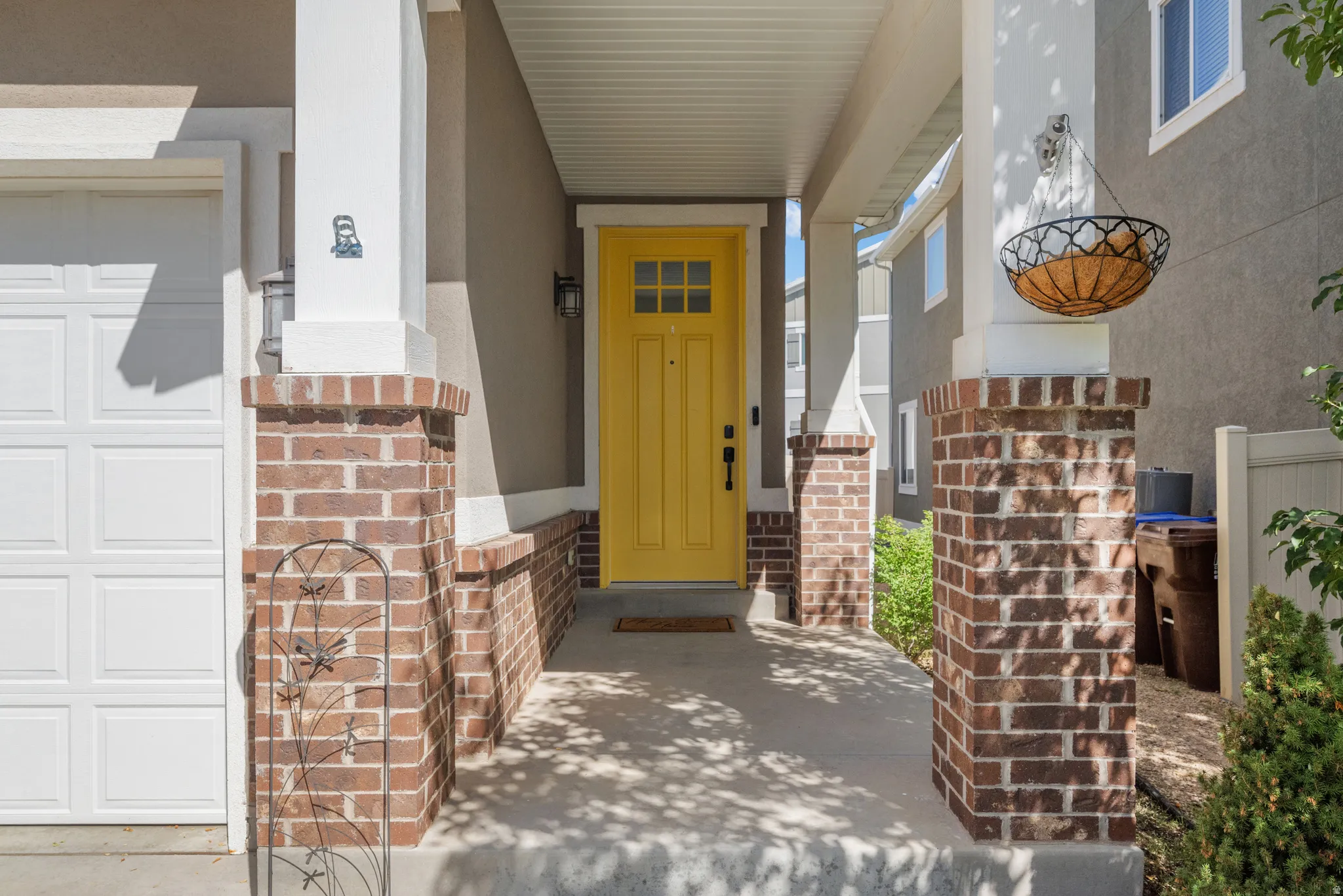 Covered porch with brick and white column accents, bright yellow front door with window panels, exterior wall lantern, and concrete walkway