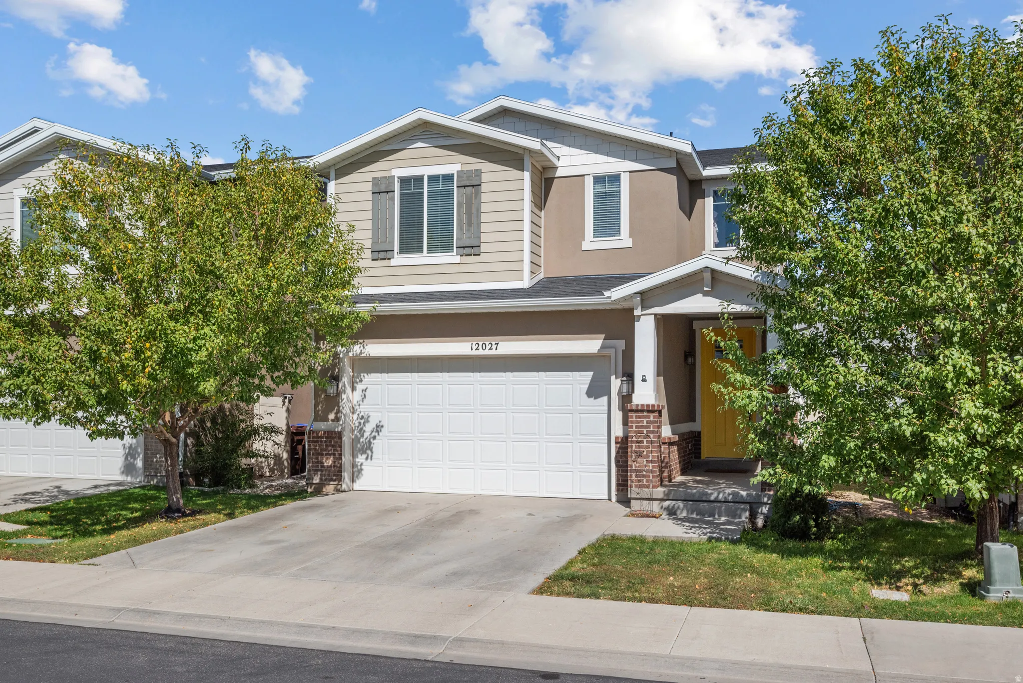 Two-story townhome with a light-colored stucco and siding exterior, an attached garage, a covered front porch with brick accents, and a bright yellow front door