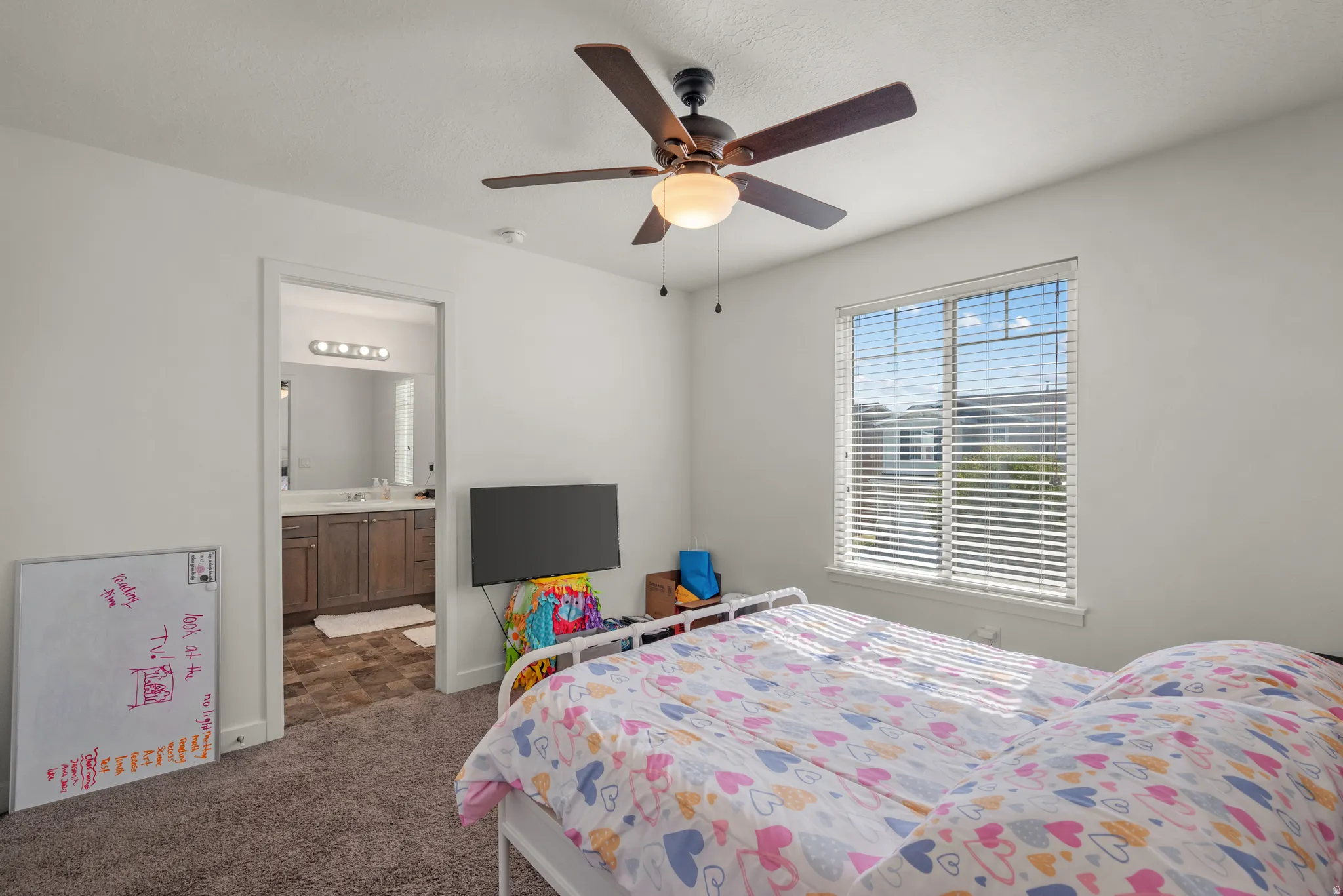 Carpeted room with a ceiling fan, window with blinds, and an attached bathroom featuring a vanity with a mirror and overhead lighting