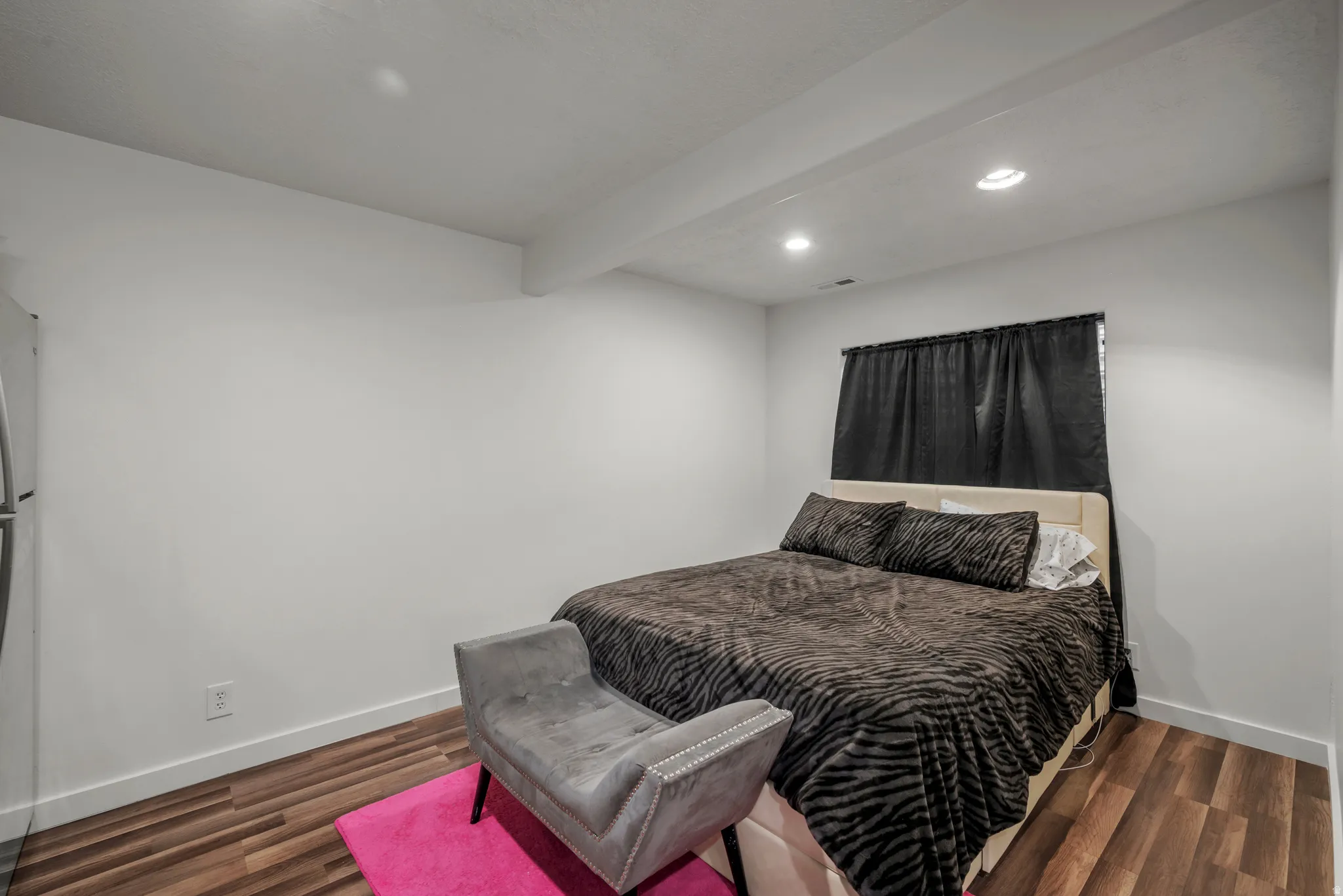 Recessed ceiling lighting, wood-finish flooring, white walls, and a window with dark drapery
