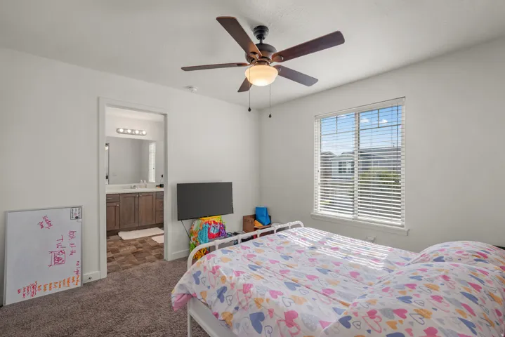 Carpeted room with a ceiling fan, window with blinds, and an attached bathroom featuring a vanity with a mirror and overhead lighting