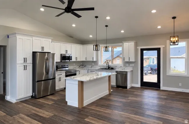 Vaulted ceiling kitchen featuring wood-finish flooring, white cabinetry, stainless steel appliances, and an island with a stone countertop