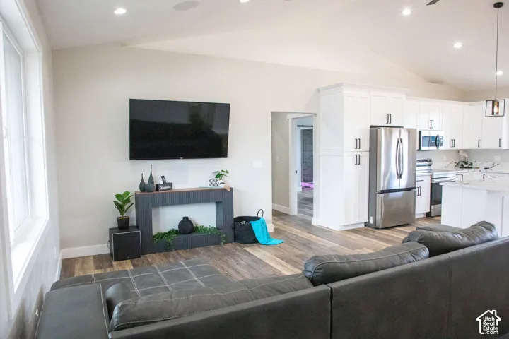 Open-concept living space featuring vaulted ceilings, recessed lighting, wood-finish flooring, white kitchen cabinetry, and stainless steel appliances