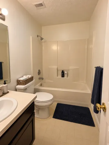 Full bathroom featuring a shower-tub combination with integrated shelving, a single vanity with an integrated sink, a toilet, and light-toned flooring