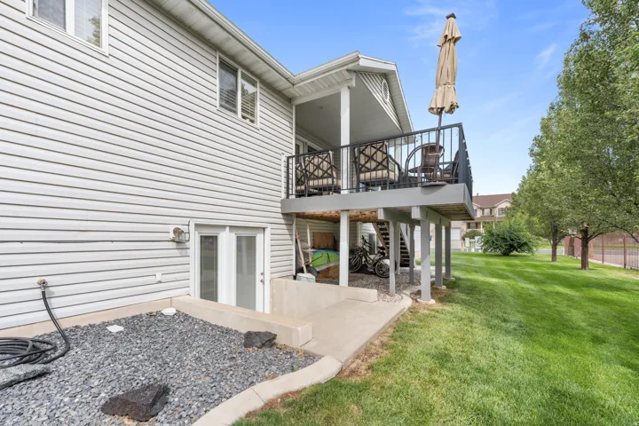 Elevated deck with black metal railing, concrete patio, double glass doors, and a spacious grassy area with mature trees