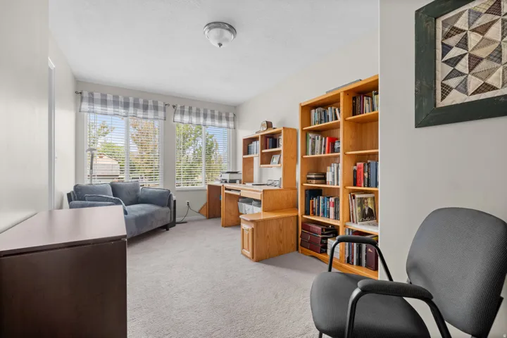 Carpeted bedroom used as office, featuring multiple windows with blinds, a built-in wood desk, and a large wood bookshelf