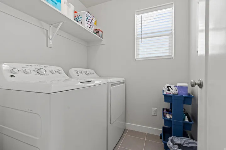 Laundry area featuring white appliances, a window with blinds, upper shelving, and tiled flooring