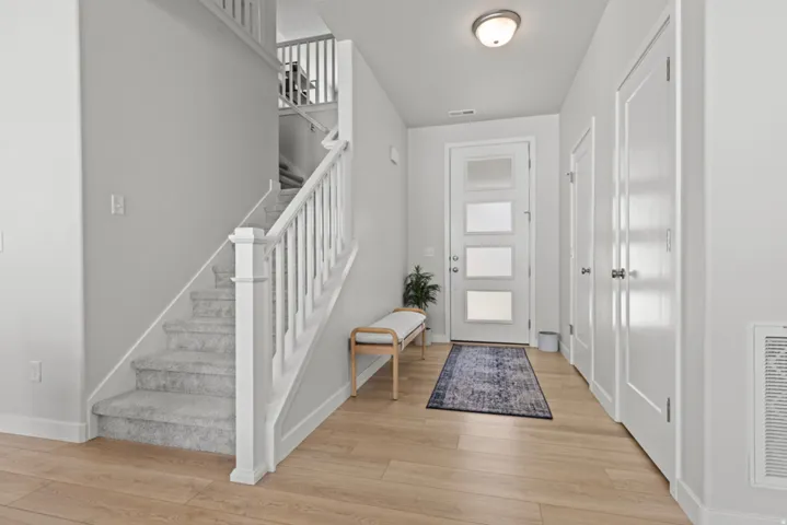 Light wood-finish flooring, staircase with carpeted treads, white balustrade, recessed ceiling light, and a contemporary front door with multiple frosted glass panels