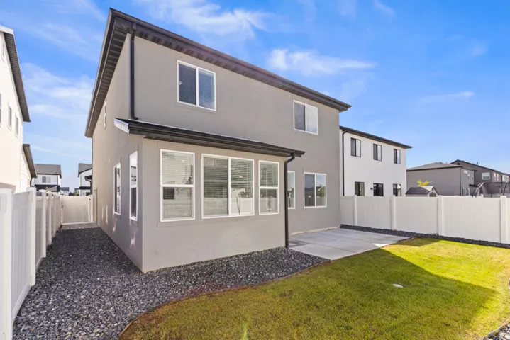 Exterior featuring stucco siding, multiple windows with white trim, a concrete patio, a grassy lawn area, and white vinyl fencing