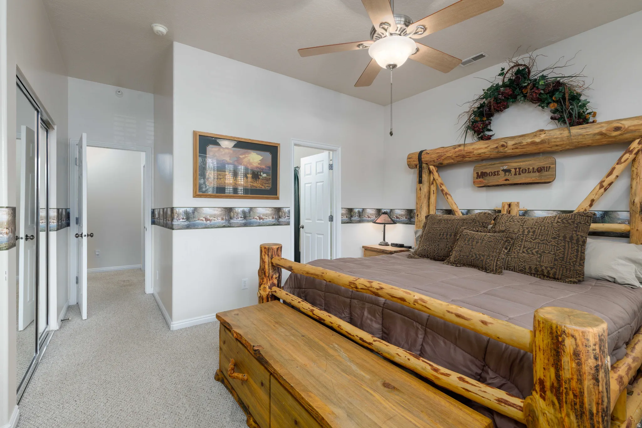 Primary Bedroom Suite with Rustic Log Bed Frame, Ceiling Fan with Integrated Lighting, Ensuite Bathroom Access, Carpeted Flooring, and Decorative Wooden Beam Accents Above Headboard