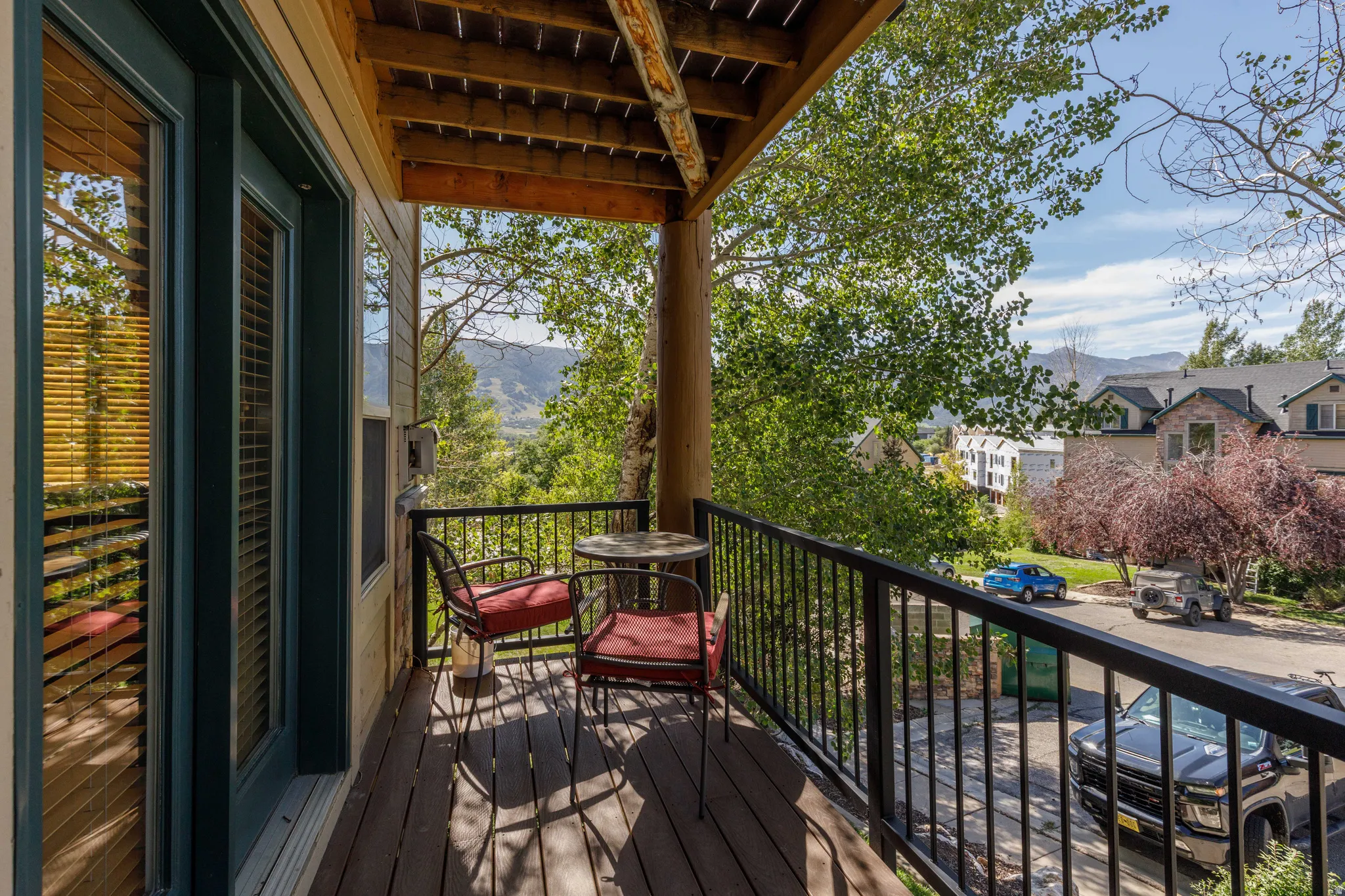 Spacious wood-plank balcony with black metal railings, offering mountain and neighborhood scenery, featuring a prominent wood beam ceiling