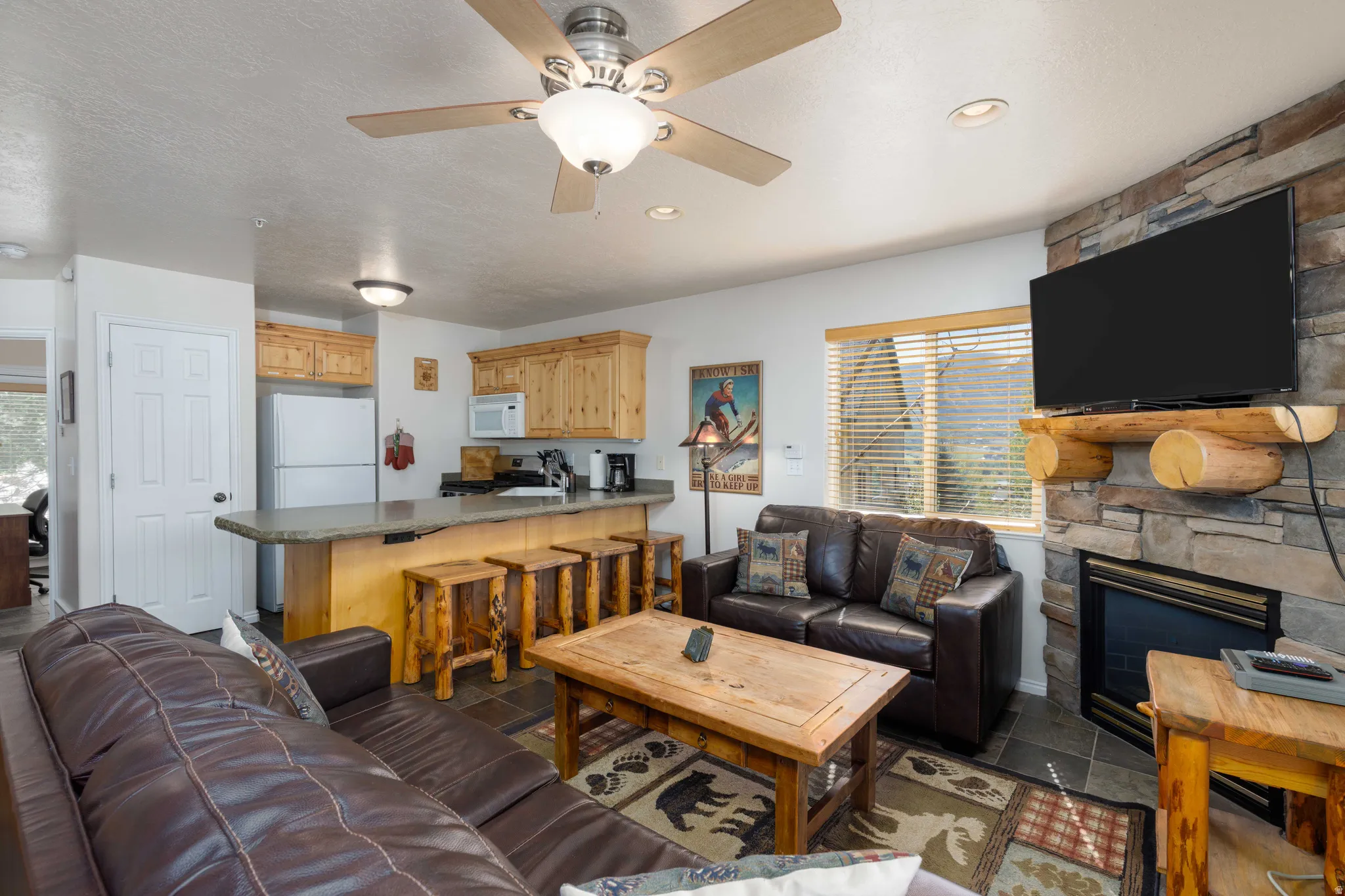 Open-concept living space featuring a wood-finish bar with seating, leather sectional sofa, wood coffee table, flat-screen television mounted above a fireplace with stone surround, ceiling fan with integrated lighting, and wood cabinetry throughout