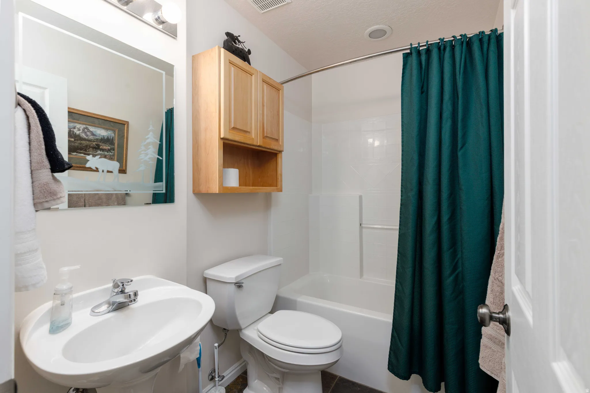 Bright bathroom featuring a round pedestal sink with chrome faucet, white subway tile surround, wooden wall-mounted cabinetry with open shelving, ceramic toilet, and a tub/shower combination with teal shower curtain and white tile surround