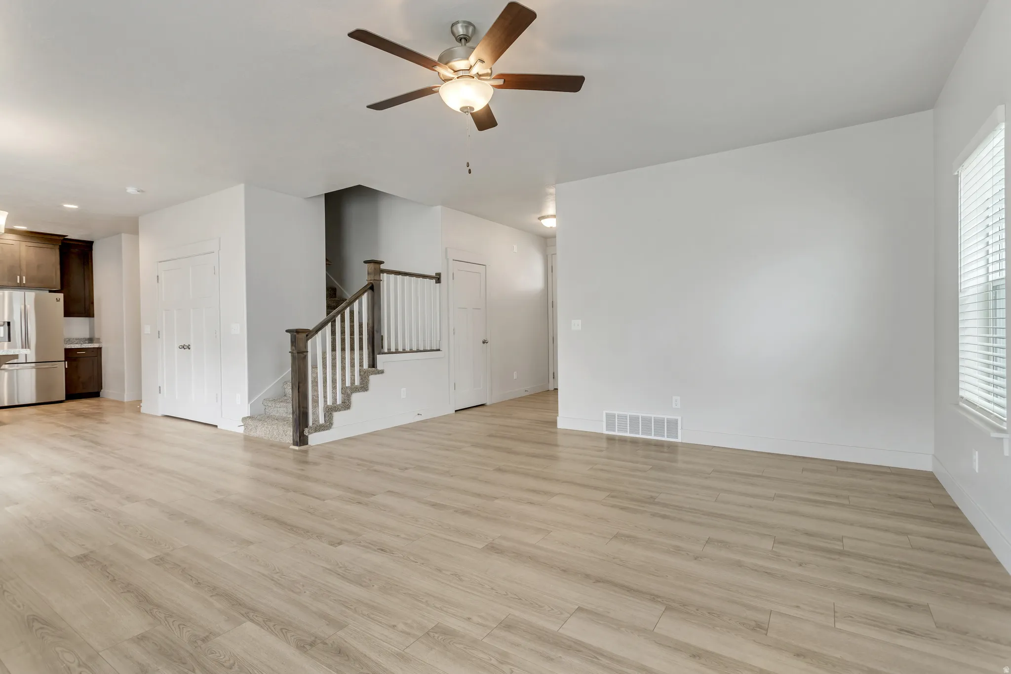 Wood-finish flooring, staircase with wood newel post and white balusters, overhead ceiling fan with lighting, white interior doors, and a window with blinds