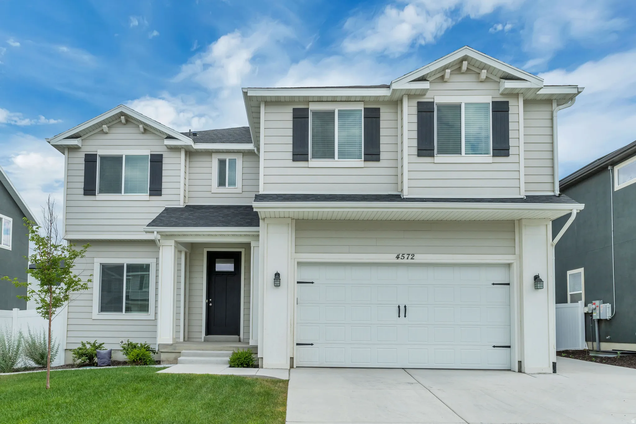Two-story residence featuring light gray siding, dark shutters, an attached garage, a covered entry, and a concrete driveway