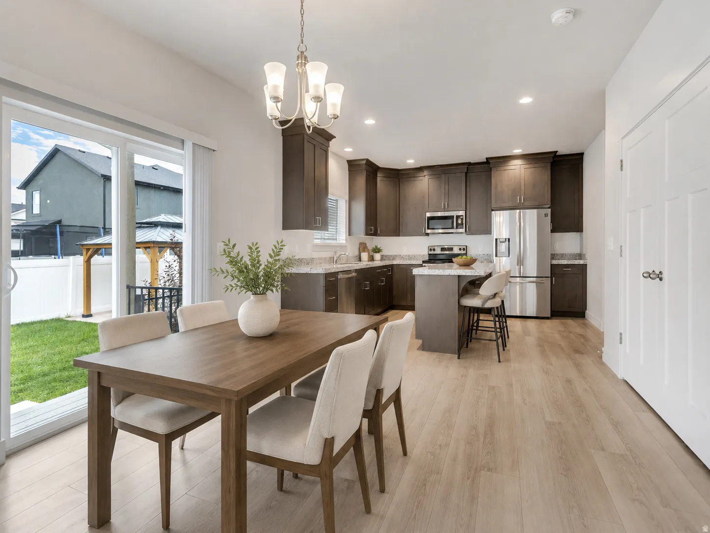 Open-concept living space with light wood-finish flooring, a modern kitchen featuring stainless steel appliances, dark wood cabinetry, and light countertops, a dining area with an overhead chandelier, and sliding glass doors leading to a backyard with a w