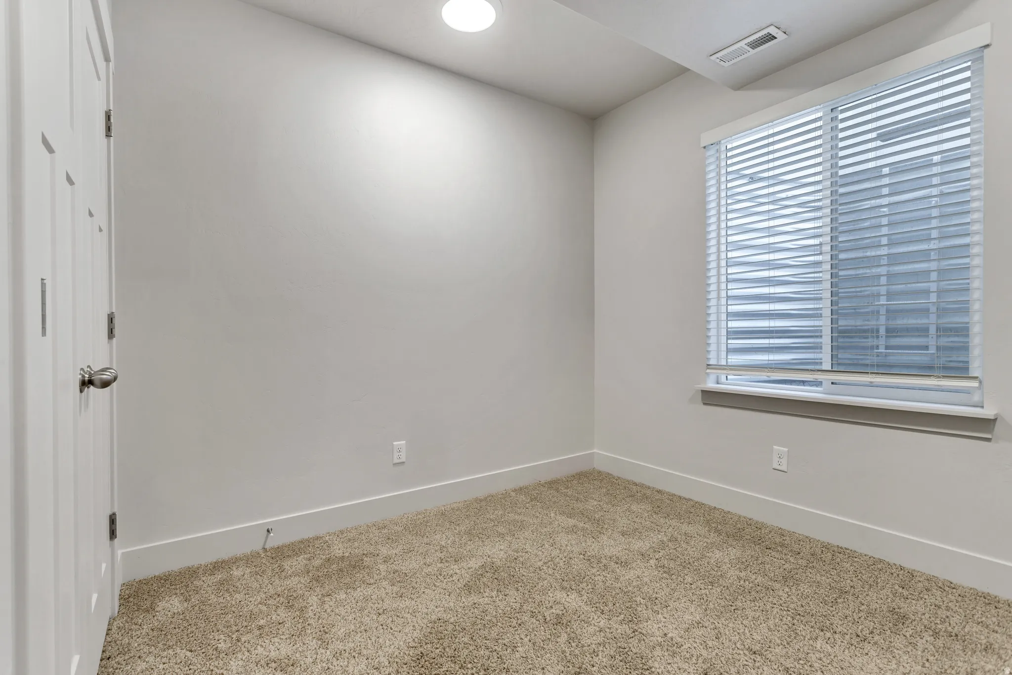 Neutral tone wall finish, light-colored carpet flooring, recessed ceiling light, white window blinds, and white interior door
