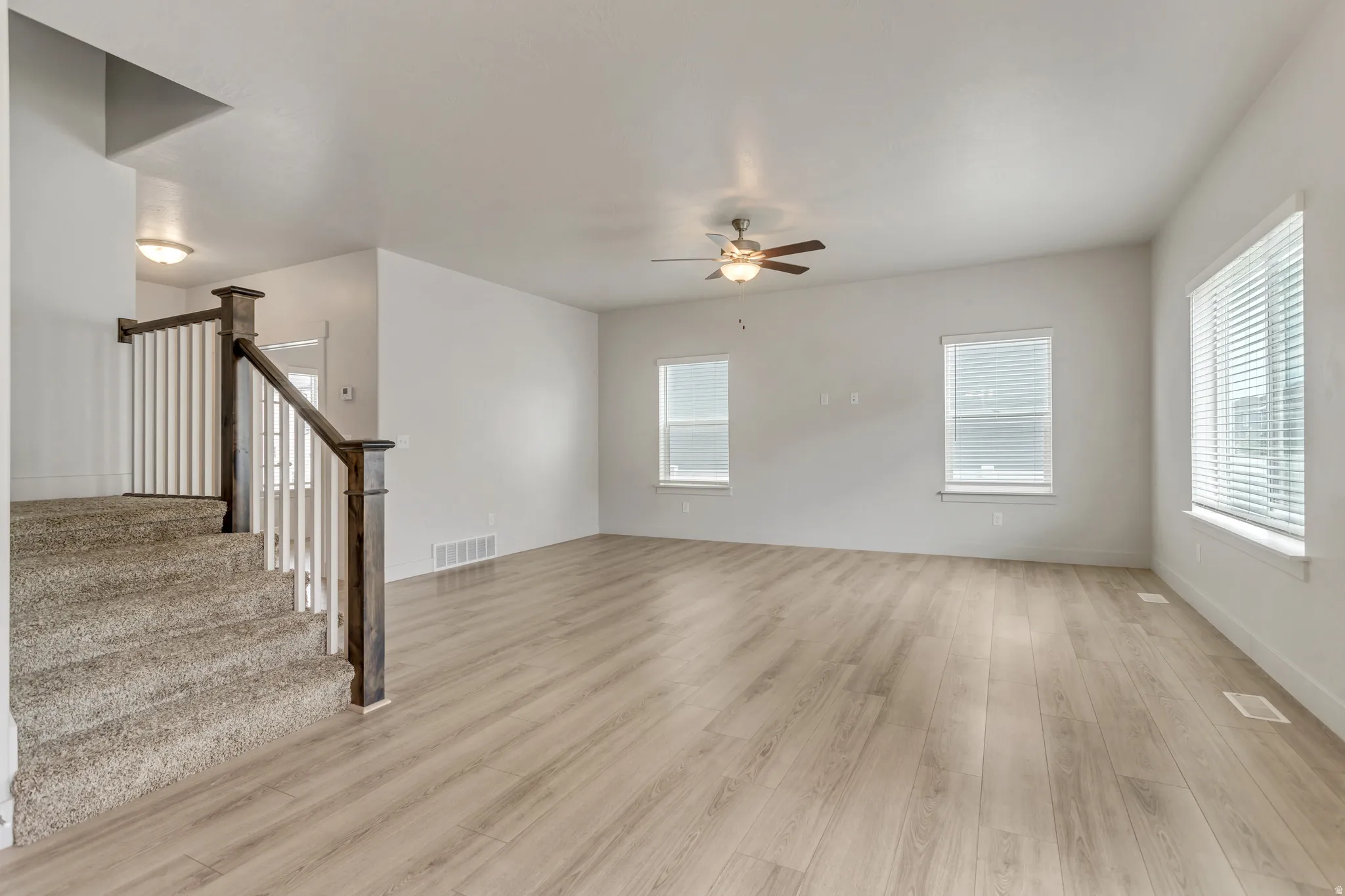 Spacious living area featuring wood-finish flooring, two large windows with blinds, a ceiling fan with integrated lighting, and a carpeted staircase with a wood and white spindle railing