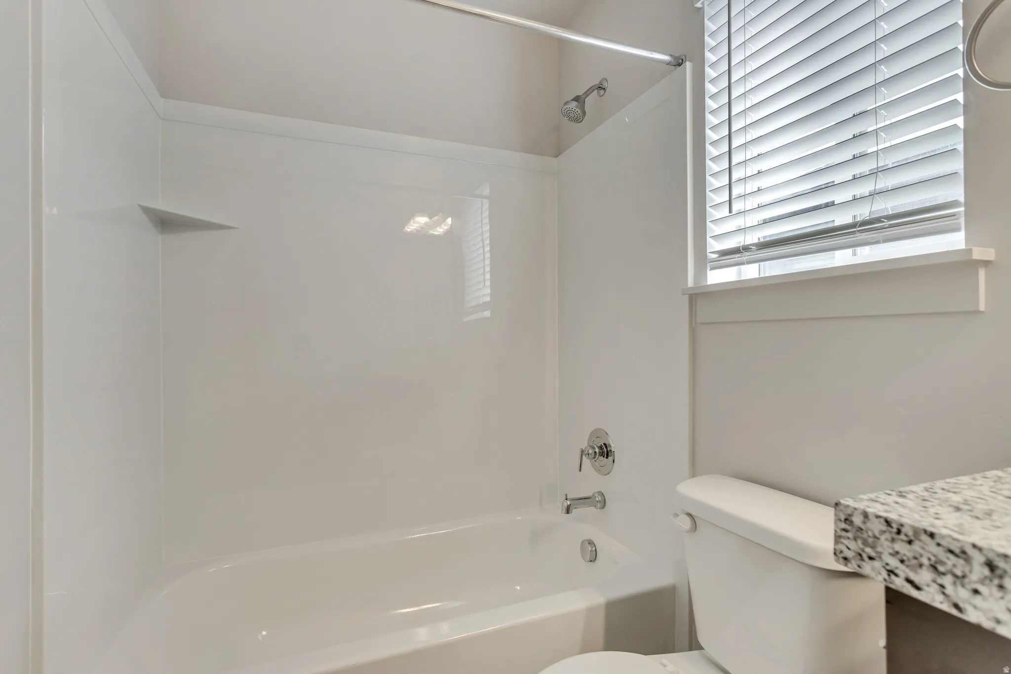 White bathtub with integrated shelving, chrome fixtures, a window with horizontal blinds, and a speckled countertop