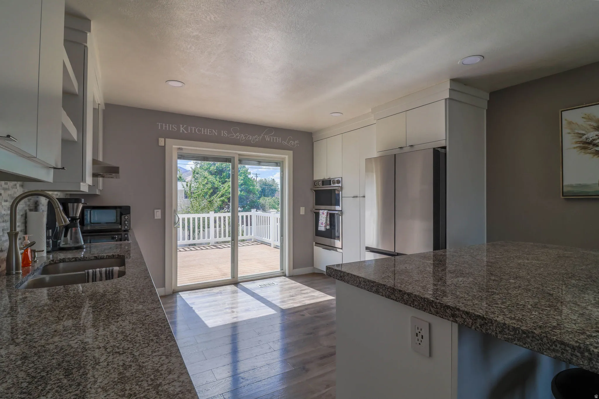 Kitchen featuring granite countertops, stainless steel appliances, white cabinetry, wood-finish flooring, and a sliding glass door leading to a deck