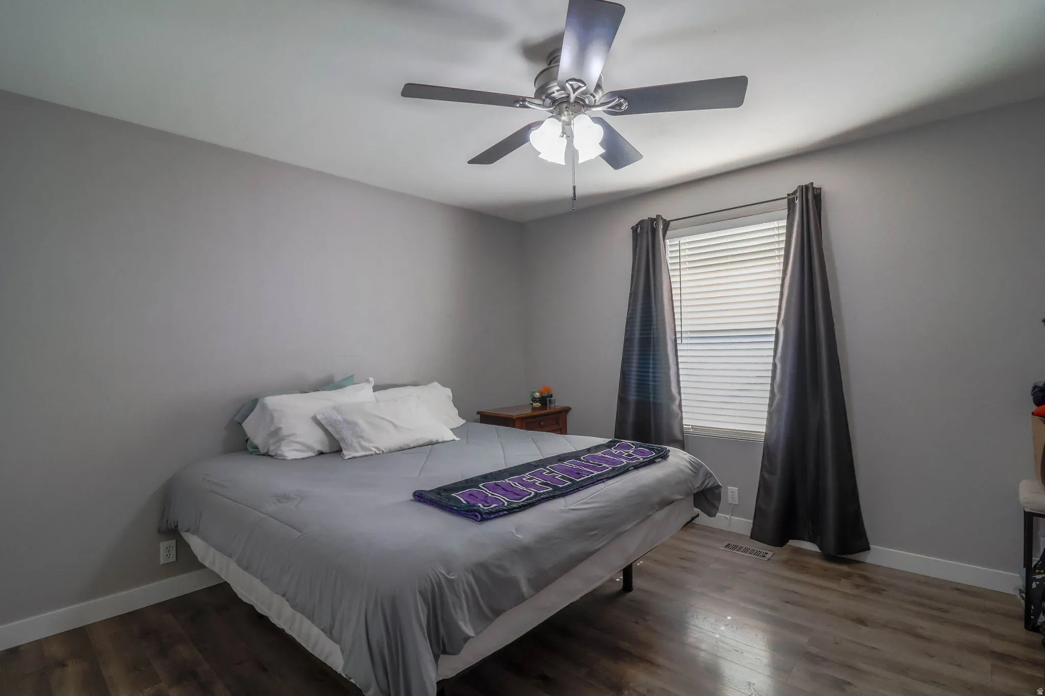 Wood-finish flooring, neutral wall paint, ceiling fan with integrated lighting, window with horizontal blinds, and dark drapery panels