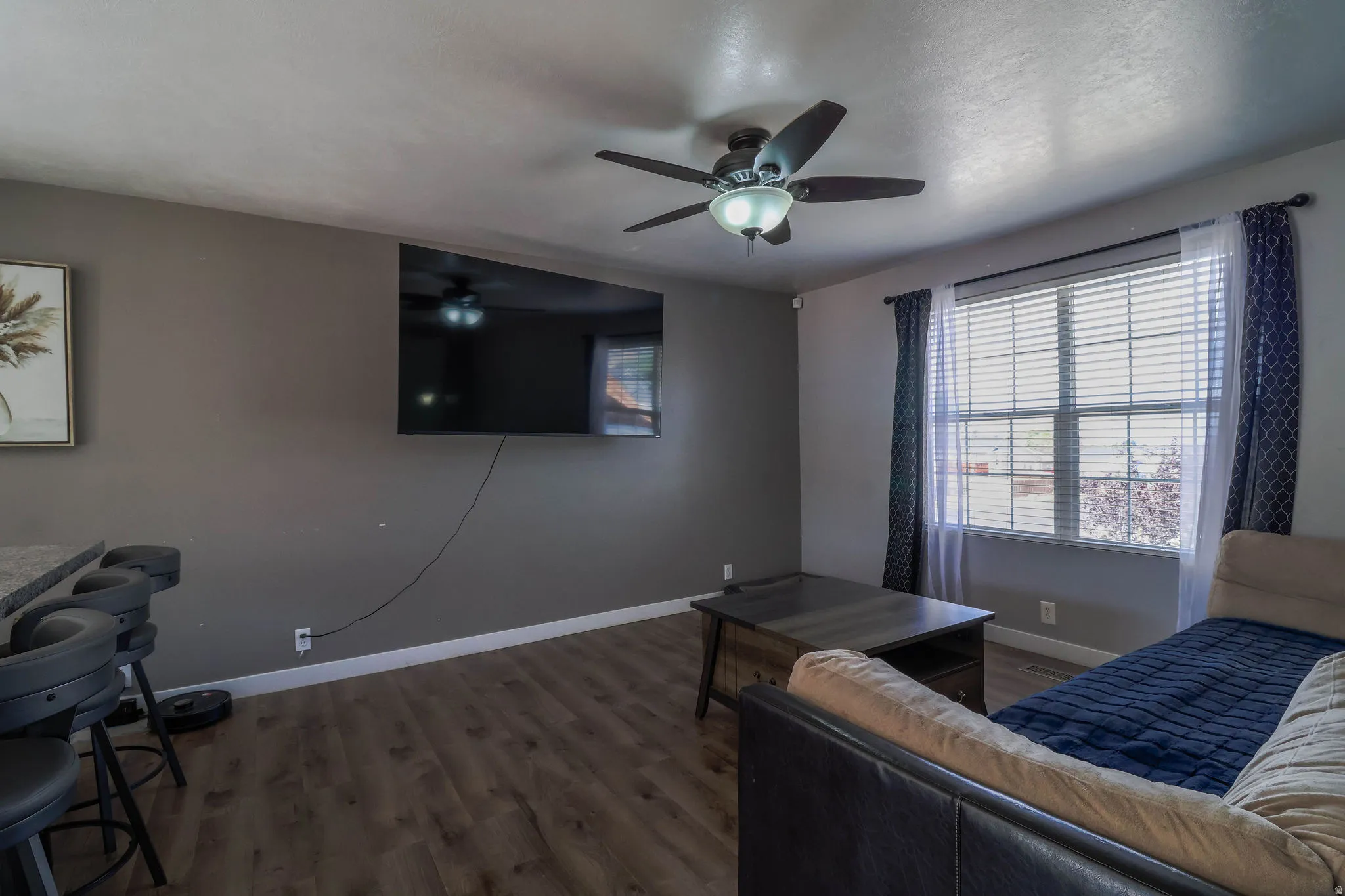 Spacious room featuring wood-finish flooring, a ceiling fan with integrated lighting, a large window with blinds, and a built-in breakfast bar with stone-look countertop