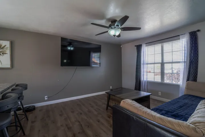 Spacious room featuring wood-finish flooring, a ceiling fan with integrated lighting, a large window with blinds, and a built-in breakfast bar with stone-look countertop
