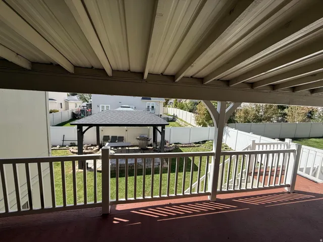 Covered outdoor deck with white railing, overlooking a fenced backyard with a gazebo, fire pit, and stone patio