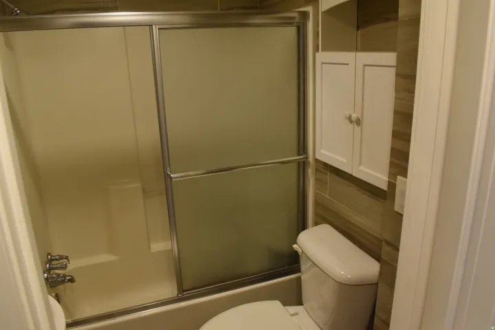 Shower-tub combination with sliding glass doors, white built-in wall cabinet, toilet, and tiled wall