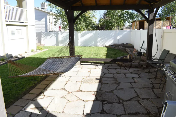 Expansive backyard featuring a stone patio, covered gazebo structure, manicured lawn, and a white privacy fence