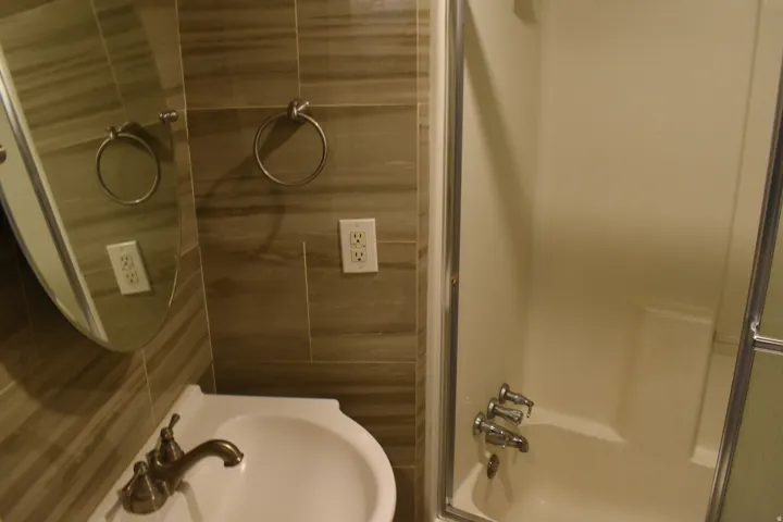 Full bathroom featuring a pedestal sink with a bronze-finish faucet, a rounded mirror, a tiled accent wall, and a bathtub with a shower enclosure