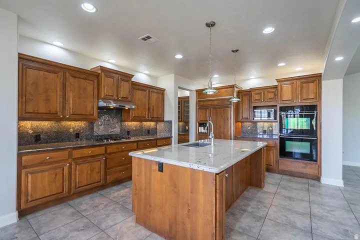 Spacious kitchen featuring wood cabinetry, granite countertops, a central island with a sink, built-in double ovens, and tile flooring